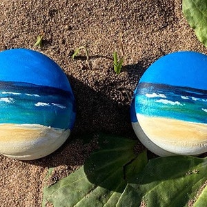 May include: Two round stones painted to look like a beach scene. The top half of each stone is painted blue to represent the sky, the middle is painted green and blue to represent the ocean, and the bottom is painted yellow to represent the sand.