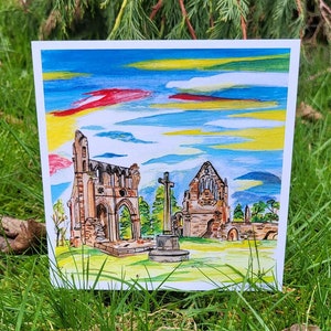Puede incluir: Una pintura de acuarela de una antigua abadía de piedra con una cruz en primer plano. El cielo es azul brillante con nubes amarillas y rojas. La abadía está en ruinas, pero la cruz sigue en pie.