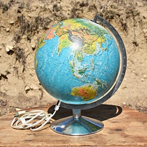 Pode incluir: Um globo vintage com um oceano azul e continentes coloridos. O globo é montado em um suporte de metal prateado e tem um anel meridiano preto. Um cabo elétrico branco está enrolado na superfície de madeira.