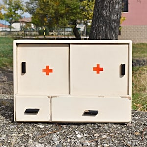 May include: A cream-coloured, vintage-style cabinet with two sliding doors and two drawers. Each door features a bright orange cross, and the cabinet has black handles. The cabinet is likely for storing medical supplies.