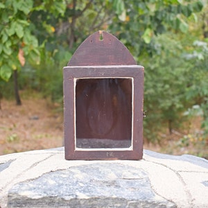 May include: A dark brown wooden shadow box with a peaked top and a rectangular opening. The frame has a white interior border. The box is displayed outdoors against a blurred green background.