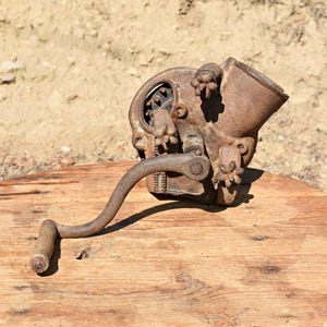 Könnte beinhalten: Eine antike, rostige Metallmühle mit gebogenem Griff und Holzgriff liegt auf einem runden Holztisch. Die Mühle hat dekorative Elemente und eine trichterförmige Oberseite. Der Hintergrund ist verschwommen und erdig.