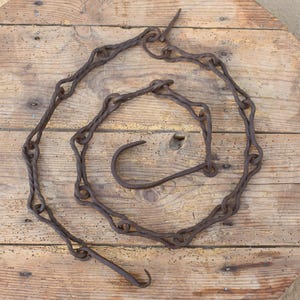 May include: A rusty brown metal chain arranged in a spiral shape. The chain has decorative links and two curved hooks at the ends. The chain is lying on a light brown wooden surface.