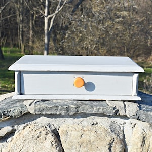 Puede incluir: Una pequeña caja de madera blanca con un solo cajón y una perilla naranja. La caja está sentada sobre una superficie de piedra.