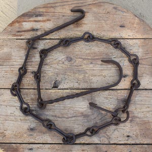 Pode incluir: Corrente de metal enferrujado com ganchos, disposta em círculo. A corrente de ferro mostra sinais de desgaste e uso. Os ganchos são curvos e parecem ser forjados à mão. Fundo de madeira.