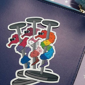 May include: A sticker of three worm-like creatures with rainbow colored bodies and googly eyes, each standing on a pole with a round base. The sticker is on a blue background.