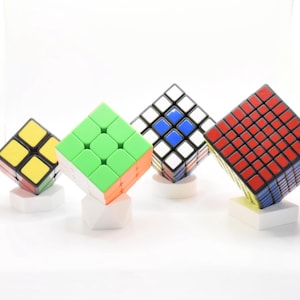 May include: Four colorful Rubik's Cubes of varying sizes are displayed on white pedestals. The cubes feature different color schemes, including yellow, green, orange, red, blue, and white. The cubes are a classic puzzle toy.