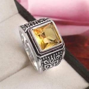 Op de afbeelding: Een zilverkleurige ring met een vierkante, gele edelsteen. De ring heeft een sierlijk ontwerp rond de edelsteen en op de band. De ring wordt gepresenteerd in een donkergroene sieradendoos.