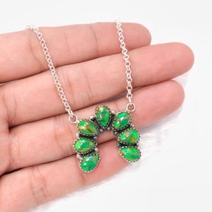 Puede incluir: Un collar de plata con un colgante en forma de herradura adornado con piedras preciosas verdes vibrantes. Las piedras tienen una apariencia texturizada única, y el collar se sostiene en una mano sobre un fondo blanco. La cadena es delicada y plateada.