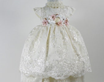 Puede incluir: Un vestido de bautizo color marfil con superposiciones de encaje floral intrincado, una falda de varias capas y un gorro a juego. La cintura está adornada con un grupo de flores de tela rosa suave y blanco, con delicados detalles perlados.