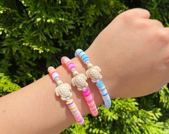 May include: Three colourful beaded bracelets with a turtle charm on each. The bracelets are pink, blue and yellow with white stripes.