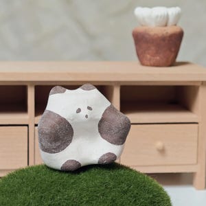 Könnte beinhalten: Handgefertigte Keramikskulptur eines stilisierten Tieres, weiß mit braunen Flecken. Sie steht auf grünem Moos, mit einem Miniatur-Holzschrank und einem kleinen braunen Topf mit weißen Blumen im Hintergrund. Ein charmantes Dekorationsstück.