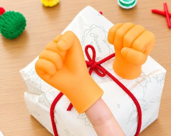 Op de afbeelding: Twee oranje vuistvormige vingerpoppetjes van flexibel materiaal. Eén poppetje zit op een vinger, terwijl de andere op een witte vierkante geschenkdoos staat die is omwikkeld met rood touw. De doos heeft een bloemmotief en het woord Princess.