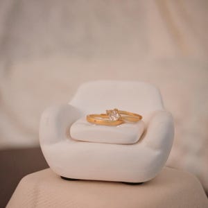 May include: Two gold rings with a square-cut diamond and a star detail rest on a small white chair. The rings are set against a soft, neutral background, creating a minimalist and elegant aesthetic.