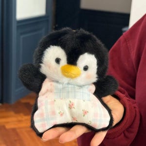 May include: A plush penguin toy with black and white fur, a yellow beak, and rosy cheeks. The penguin wears a pink and blue patterned dress with floral accents. The toy is designed to be held.