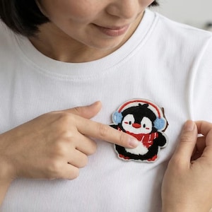 Pode incluir: Uma camiseta branca com um patch bordado colorido de um pinguim de desenho animado. O pinguim tem um corpo preto, barriga branca, cachecol vermelho e protetores de ouvido azuis. Um dedo aponta para o patch.
