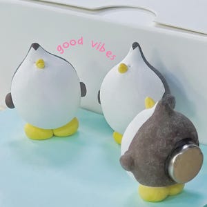 May include: Three penguin figurines with white bodies, black backs, and yellow feet. The text "good vibes" is written in pink above the penguins. The penguins have small brown ears and yellow beaks.