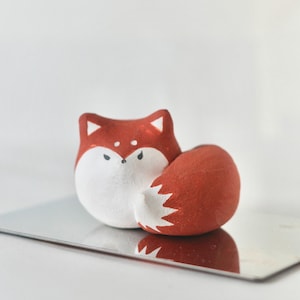 May include: A handcrafted fox figurine with a red body, white face, and a fluffy white-tipped tail. The fox is sitting on a reflective silver surface. The fox has black eyes and is a decorative item.