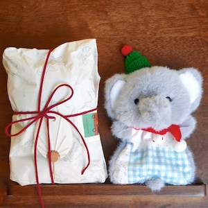 May include: A rectangular gift package wrapped in white paper with a faint floral pattern, tied with red string and a small white fan ornament, next to a plush grey elephant toy wearing a green knit hat and a red scarf over a blue checkered cloth.