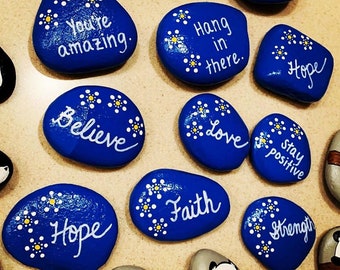 May include: A collection of painted rocks with inspirational messages, including "You're amazing", "Believe", "Hope", "Love", "Faith", "Hang in there", "Stay positive", and "Strength". The rocks are painted blue with white lettering and yellow flower accents.
