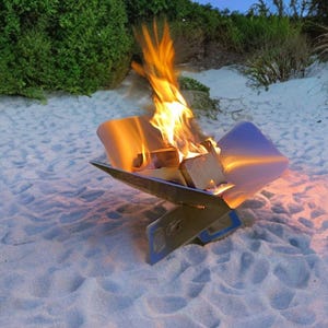 May include: A stainless steel fire pit with a blue trim is set up on a sandy beach. The fire pit is lit with a bright orange flame, and there are logs inside. The background features green bushes and a blue sky.