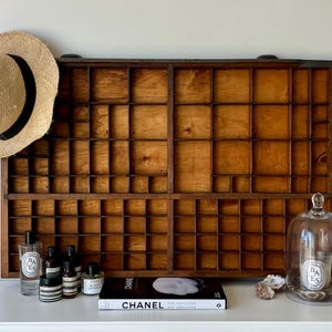 May include: A vintage wooden printer's tray with multiple compartments, a straw hat with a black band, and various apothecary bottles. A glass cloche with a candle and a book with the word "CHANEL" are also present.