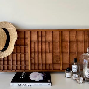 May include: A rectangular wooden printer's tray with multiple small compartments, a straw hat with a black band, a Chanel book, two glass perfume bottles, a glass cloche covering a candle, and two small white crystal clusters arranged together.