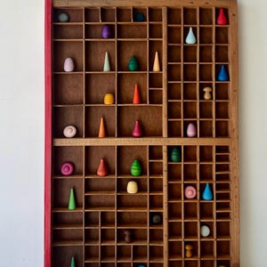 May include: A wooden display case with many small compartments, each containing a colourful wooden toy. The toys are various shapes, including cones, eggs, and mushroom-like forms, painted in shades of red, green, blue, yellow, and purple.