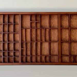 May include: A rectangular wooden printer's tray featuring a natural brown wood grain finish. The tray is divided into numerous small, varied compartments of different sizes and shapes, organized into three distinct vertical sections.