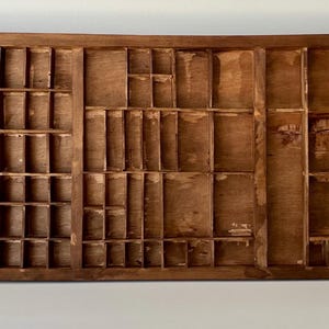 May include: A rectangular wooden printer's tray featuring a grid of numerous small, open compartments of varying sizes. The wood has a medium brown finish with visible grain and signs of wear, organized into three distinct vertical sections.
