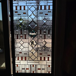 May include: A stained glass window with a geometric pattern in shades of brown, white, and gray. The window is set in a black frame and is illuminated by sunlight.