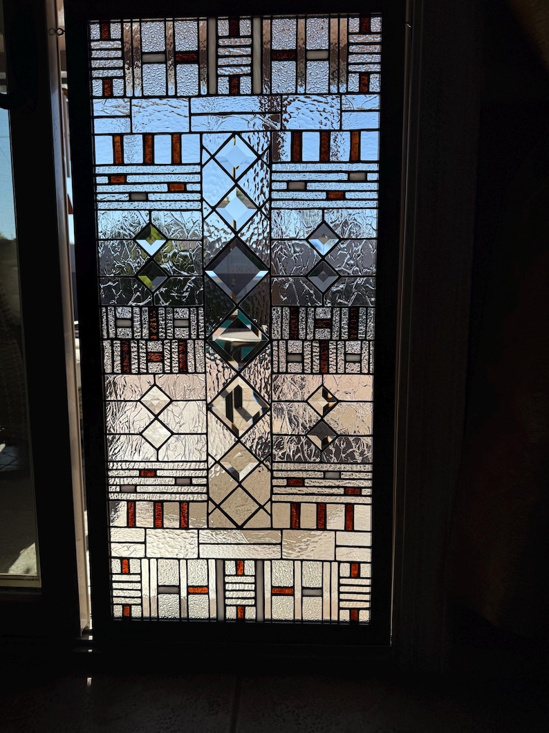 May include: A stained glass window with a geometric pattern in shades of brown, white, and gray. The window is set in a black frame and is illuminated by sunlight.