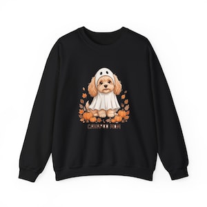 Puede incluir: Una sudadera negra de manga larga con un gráfico de un perro Cavapoo marrón claro vestido con un disfraz de fantasma blanco. El perro está rodeado de calabazas naranjas y hojas de otoño marrones, con el texto CAVAPOO MOM impreso en marrón.