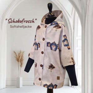 Könnte beinhalten: Hellfarbene Softshelljacke mit Kapuze, mit einem sich wiederholenden Muster aus Frosch- und Zauberer-Illustrationen. Die Jacke hat dunkelblaue Ärmel und braune Knöpfe. Der Text "Schokofrosch Softshelljacke" ist oben links aufgedruckt.