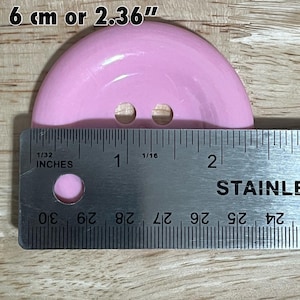 May include: A pink plastic button with two holes, measuring 2.36 inches in diameter. The button is being measured with a ruler.
