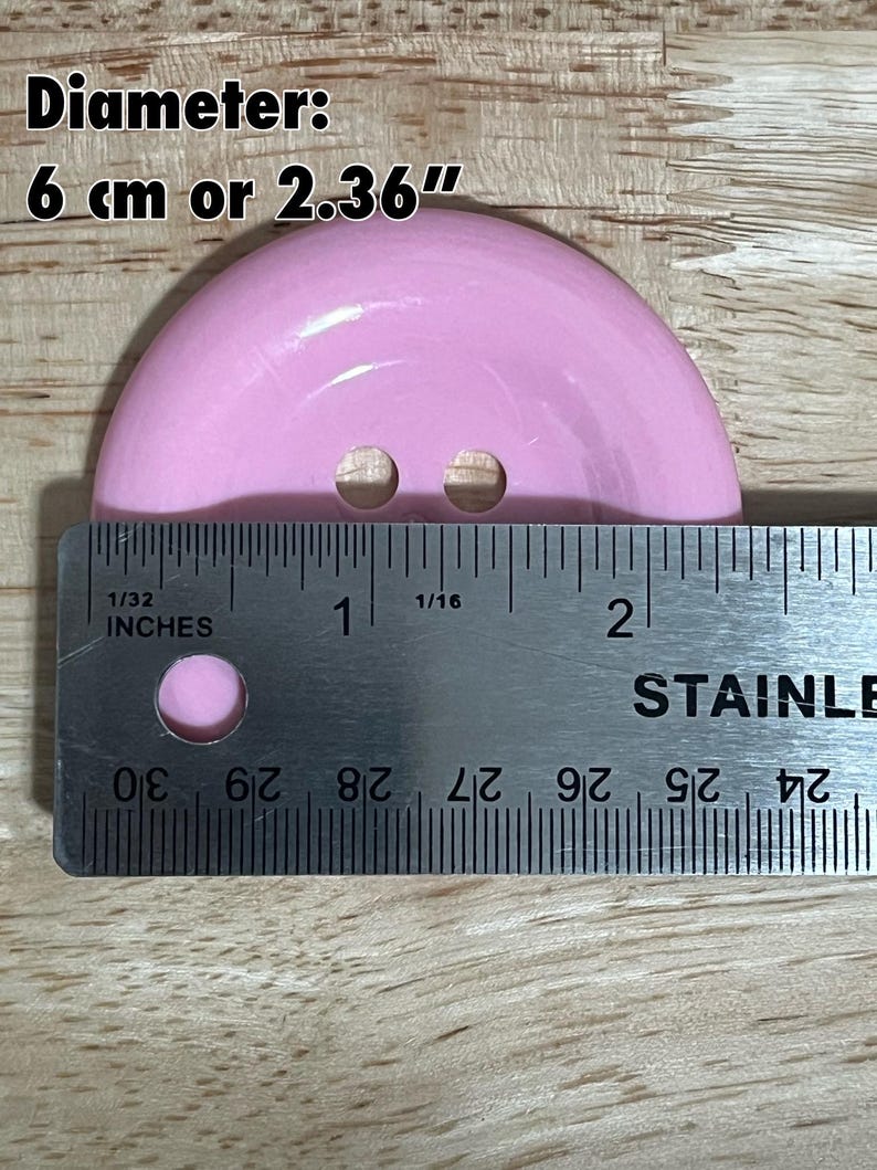 May include: A pink plastic button with two holes, measuring 2.36 inches in diameter. The button is being measured with a ruler.