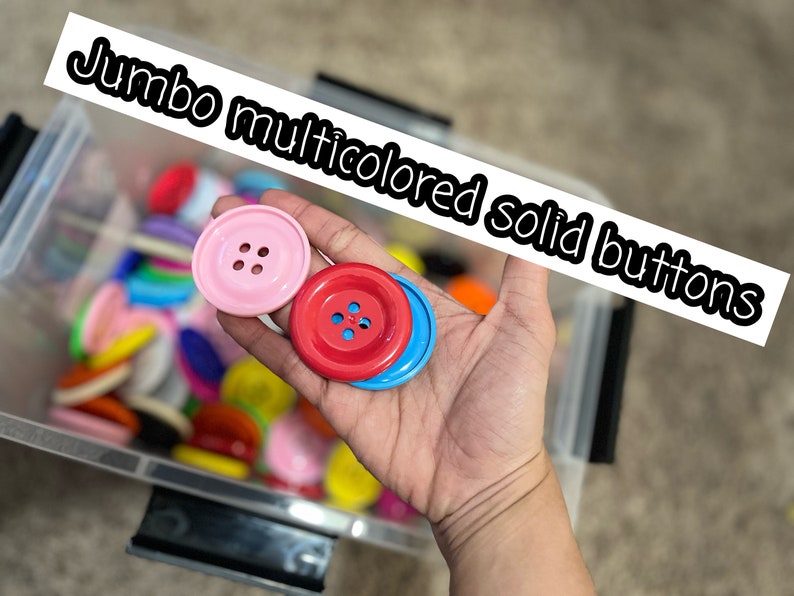 May include: A hand holding three large, colorful buttons. The buttons are pink, red, and blue. The buttons have four holes each. The buttons are in a container with many other buttons of various colors.