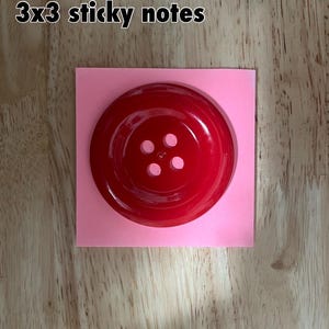 May include: A large, red, plastic button with four holes. The button is sitting on a pink square.