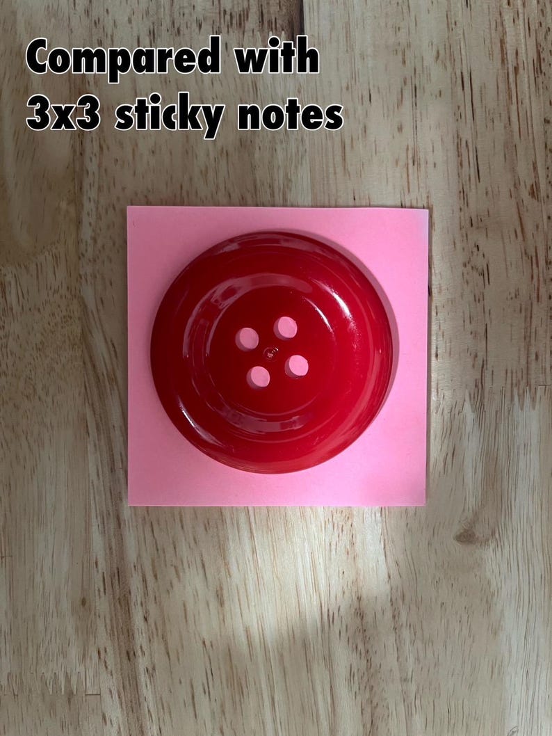 May include: A large, red, plastic button with four holes. The button is sitting on a pink square.