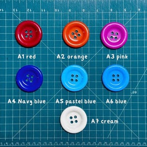 May include: Seven different colored buttons with four holes each. The buttons are arranged in three rows on a blue cutting mat. The buttons are red, orange, pink, navy blue, pastel blue, blue, and cream.