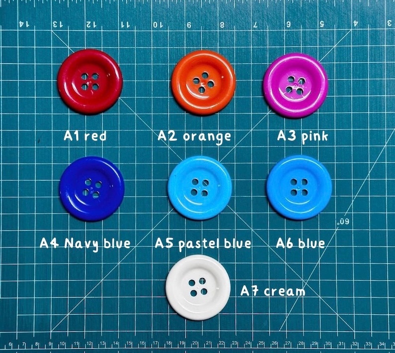 May include: Seven different colored buttons with four holes each. The buttons are arranged in three rows on a blue cutting mat. The buttons are red, orange, pink, navy blue, pastel blue, blue, and cream.