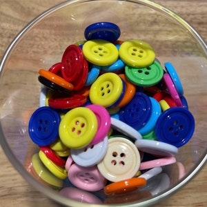 May include: A glass bowl filled with colorful plastic buttons in various shapes and sizes. The buttons are red, yellow, blue, green, pink, and white.
