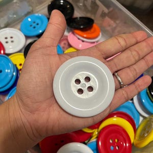 May include: A collection of colorful plastic buttons in various sizes and colors, including red, blue, yellow, white, black, and orange. The buttons have four holes each and are arranged in a container.