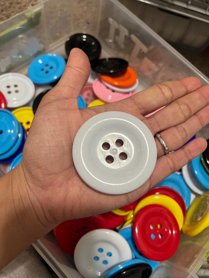 May include: A collection of colorful plastic buttons in various sizes and colors, including red, blue, yellow, white, black, and orange. The buttons have four holes each and are arranged in a container.