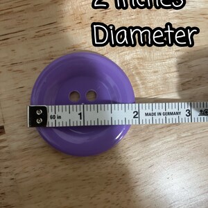 May include: A purple plastic button with two holes, measuring 2 inches in diameter. The button is on a wooden surface with a measuring tape.