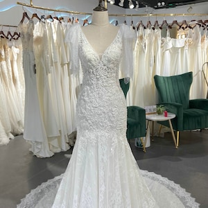 May include: A white lace wedding dress with a V-neckline and short sleeves. The dress has a fitted bodice and a flowing skirt. The dress is displayed on a mannequin in a bridal shop.