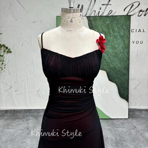 K&ouml;nnte beinhalten: Ein schwarzes Netzkleid mit einem roten Blumenakzent am Tr&auml;ger. Das Kleid hat ein tailliertes Oberteil und einen flie&szlig;enden Rock. Das Kleid befindet sich auf einer Schaufensterpuppe.