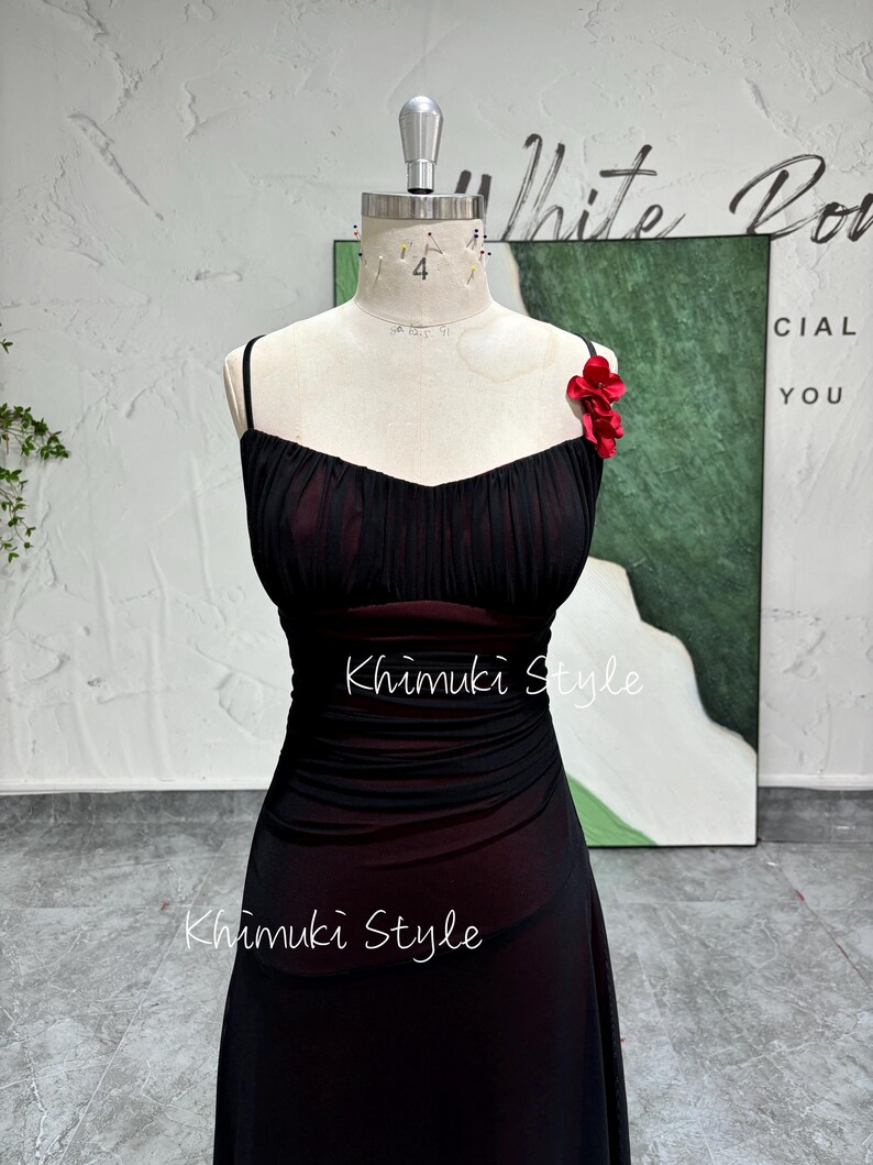 K&ouml;nnte beinhalten: Ein schwarzes Netzkleid mit einem roten Blumenakzent am Tr&auml;ger. Das Kleid hat ein tailliertes Oberteil und einen flie&szlig;enden Rock. Das Kleid befindet sich auf einer Schaufensterpuppe.