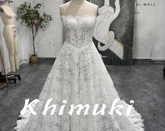 May include: A strapless white lace wedding dress on a mannequin. The dress features intricate floral lace detailing and a full skirt. The text "Khimuki Style" is displayed on the front of the dress. The mannequin is set against a neutral background.