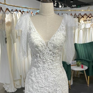 May include: A white wedding dress with a V-neckline and a fitted bodice. The dress has a lace overlay with beaded embellishments and sheer sleeves with bows. The dress is displayed on a mannequin in a store setting.